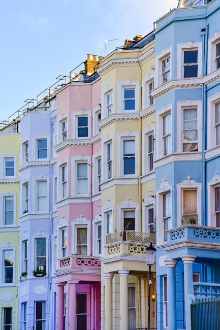 A row of colorful Victorian-style terraced houses in Notting Hill with distinctive bay windows and ornate architectural details, painted in pastel shades of blue, pink, and cream. The buildings featur