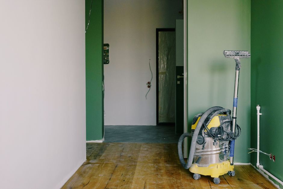 The image shows a room in the process of refurbishment cleaning, featuring a wooden floor with visible dust and debris, and a green painted wall on the right side. A professional cleaning vacuum with