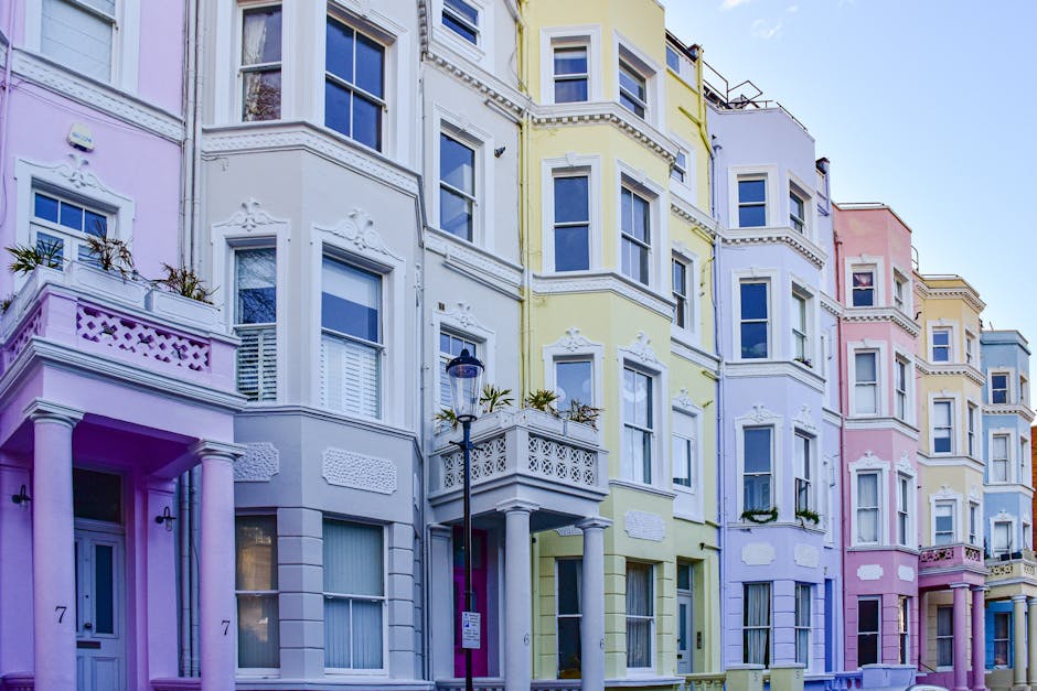 A row of Victorian-style residential buildings in Notting Hill, featuring a mix of white, light grey, and pink facades. The buildings have large, multi-pane sash windows, some with flower boxes, and d